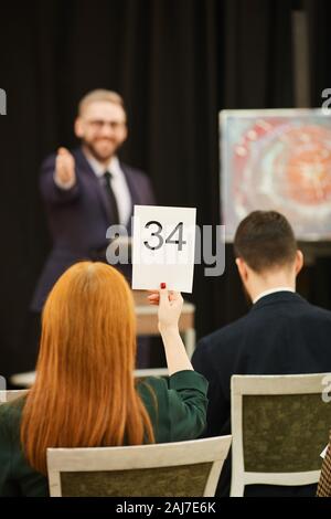 Rear view of red haired woman sitting and raising her sign and man pointing at her during auction Stock Photo