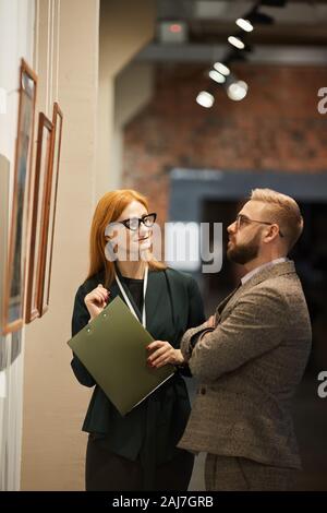 Art manager with folder and confident man standing in front of the wall with paintings and discussing it together at museum Stock Photo