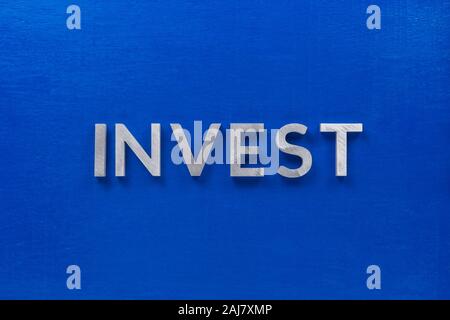 the word invest laid with silver metal characters on blue painted wooden board in central flat lay composition. Stock Photo