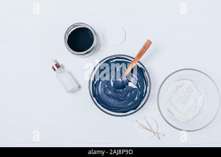 Homemade cosmetic facial mask made of clay and coal powder next to coconut oil bottle and cotton pads on white wooden table, flat lay Stock Photo