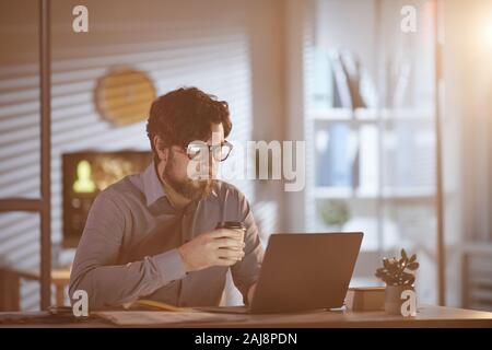 Young bearded businessman in eyeglasses sitting at his workplace drinking coffee and using laptop at office Stock Photo