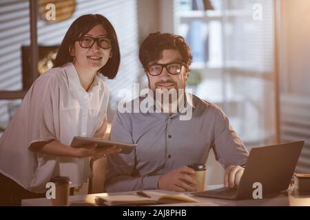 Portrait of young business people smiling at camera while working with computer and tablet pc till late at office Stock Photo
