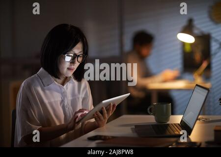 Serious Asian businesswoman in eyeglasses sitting at her workplace with laptop and using digital tablet in work at dark office Stock Photo