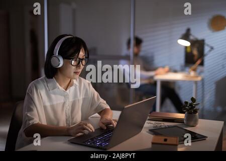 Serious Asian designer in headphones working on laptop computer at the office till late evening Stock Photo