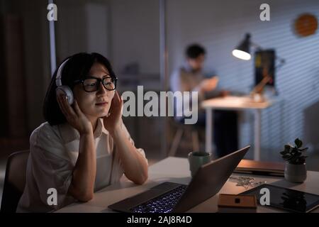 Young Asian businesswoman wearing wireless headphones and looking at monitor of laptop while sitting at her workplace at dark office Stock Photo