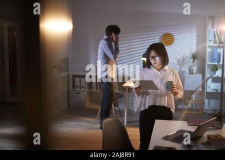 Asian businesswoman looking at tablet pc in her hands and drinking coffee with businessman talking on the phone in the background Stock Photo