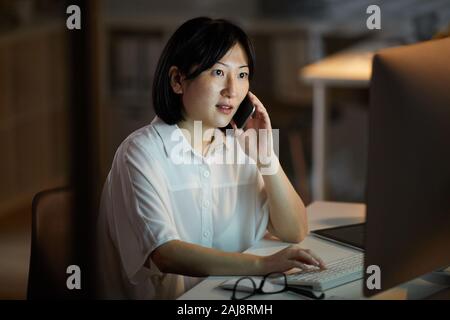 Asian young businesswoman sitting at the table typing on computer keyboard and talking on mobile phone she working till late evening Stock Photo