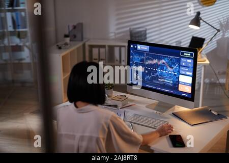 Rear view of young analyst sitting at the table in front of computer monitor with financial charts and analyzing it at office Stock Photo