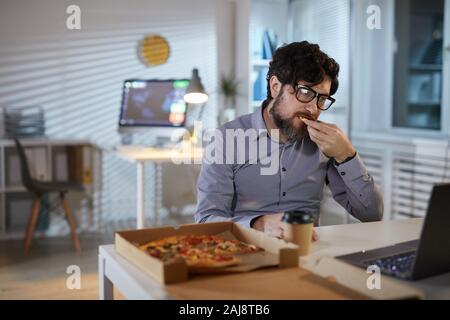 Young businessman eating pizza and looking at monitor of laptop while sitting at his workplace at office Stock Photo