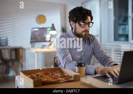 Serious bearded businessman in eyeglasses typing on laptop and eating pizza during his lunch at office Stock Photo