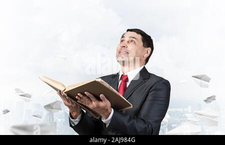 Happy businessman looking up with open book. Smiling man in suit standing on downtown background with flying paper planes. Lawyer holding big legal re Stock Photo