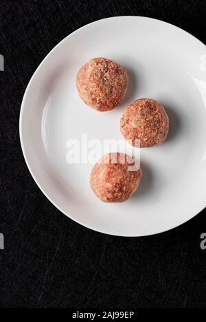 high angle view some raw homemade meatballs in a white ceramic plate, on a black stone table or countertop Stock Photo