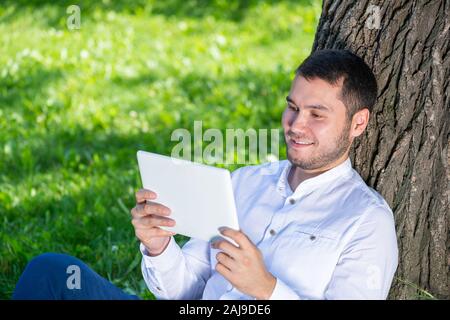 Man using tablet computer under tree in park on sunny day. Handsome guy looking at tablet screen. Happy smiling man sitting on green grass leaning aga Stock Photo