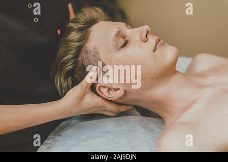 The masseur does a facial massage to a young guy. A man is massaged face on a massage table. Stock Photo