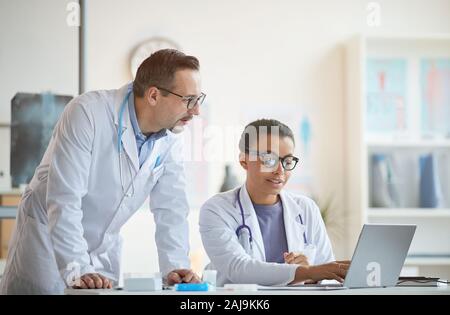 Young nurse sitting at the table and working on laptop and talking to the male doctor in white coat at modern office Stock Photo