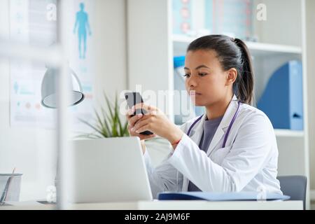 Young female doctor sitting at the table and using her mobile phone during her work at office Stock Photo