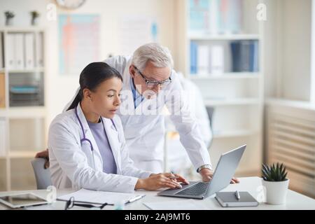 Senior doctor standing and looking at laptop computer while young nurse sitting at the table and typing on laptop at office Stock Photo