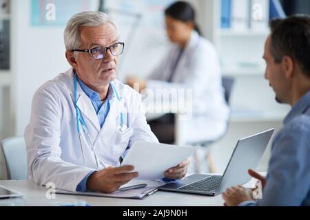 Senior doctor in eyeglasses and in white coat sitting at the table with document and explaining the treatment to his patient at office Stock Photo