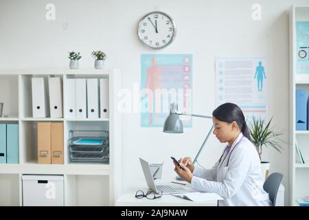 Young pediatrician sitting at her workplace in front of laptop and using mobile phone at office Stock Photo