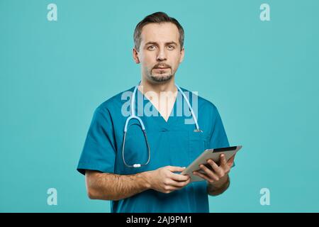 Portrait of mature surgeon in scrubs looking at camera while using digital tablet over blue background Stock Photo