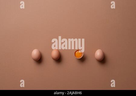 Brown eggs and one cracked egg with yolk on a brown paper background. Above view of three eggs and one half. Aligned organic eggs. Minimal image. Stock Photo