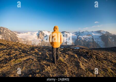 Rear view of hiker walking on mountain summit in yellow jacket. Stock Photo