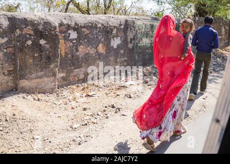 Udaipur, India - March 05 2017: Woman walking on the road with a baby in her arms. The girl wants to sleep and yawns looking around. Stock Photo