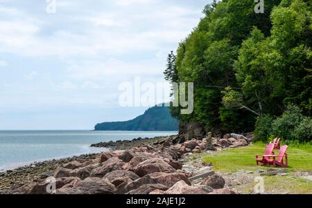 Red Adirondack chairs on Bay of Fundy shore. Shingle beach on a rugged coastline. Mixed forest in the back. Fundy National Park, New Brunswick, Canada Stock Photo