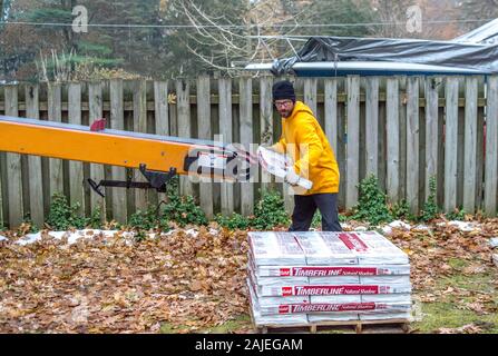 A man unloads heavy bundles of shingles from a portable conveyor belt onto a job site Stock Photo