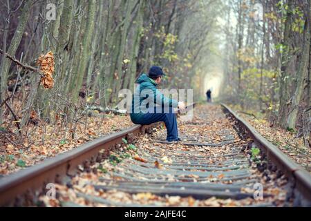 The lonely man sits on the rail of an old railway in the autumn forest Stock Photo