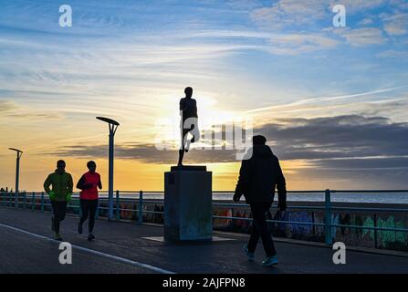 Brighton UK 4th January 2020 - Runners , walkers and cyclists enjoy a sunny but chilly morning along Brighton seafront as they pass by the statue of Olympic champion Steve Ovett at sunrise . Credit: Simon Dack / Alamy Live News Stock Photo
