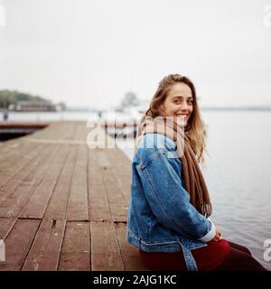 Stylish woman in red dress and blue jacket sitting on old wooden pier with a boat Stock Photo