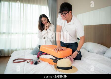 Asian man trying to fit all clothing to packing his orange suitcase before vacation and her girlfriend looking on the bed in background. in bedroom at Stock Photo