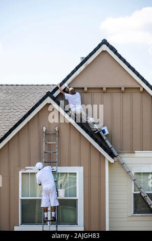 Painter man in white clothes with a protective scarf on his head paints a wooden window trim with a brush while standing on a high ladder leaning agai Stock Photo