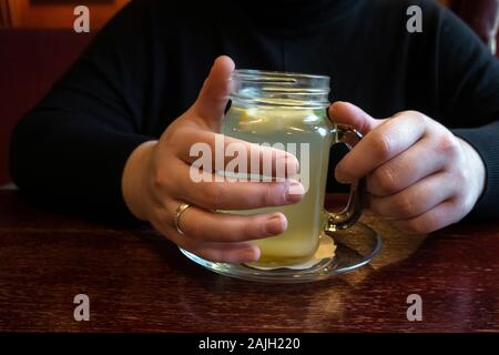 Woman drinking hot and refreshing ginger tea with slice of lemon in a restaurant. Hands holding cup of natural herbal tea on wooden table, warm winter Stock Photo
