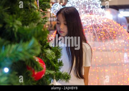 Asian teenage girl wearing a white shirt stands above the Christmas tree. Stock Photo