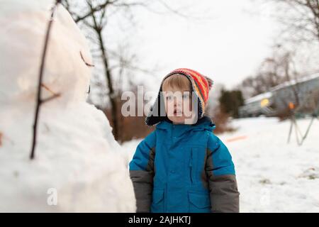 Mid shot of toddler boy wearing knit hat standing looking at snowman Stock Photo