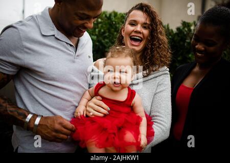 Mom Laughing & Holding Biracial Daughter as Father and Sister Look On Stock Photo