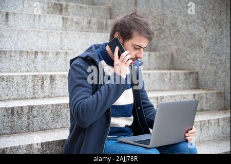Man talking on phone while working on laptop sitting in stair steps in urban scenery . Stock Photo
