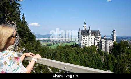 Pretty blond female tourists enjoys the view of mad King Ludwig's fairytale castle from Marienbrucke or Marien Bridge in Bavaria, Germany. Stock Photo