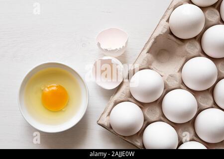 Detail of white chicken eggs in a paper tray and broken egg in a white ceramic bowl. Top view. Space for text. Stock Photo