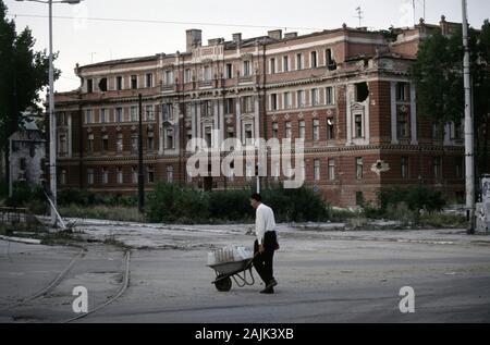 17th August 1993 During the Siege of Sarajevo: a weary man pushes a wheelbarrow loaded with containers filled with water past the bomb-damaged Supreme Court building. Stock Photo