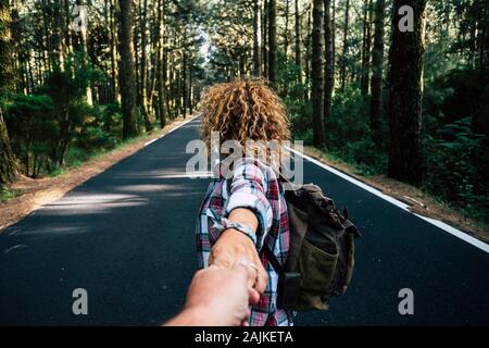 Couple travel together walking with backpack in a forest on a long asphalt road - man point of view -  holding hands with love and relationship - alte Stock Photo