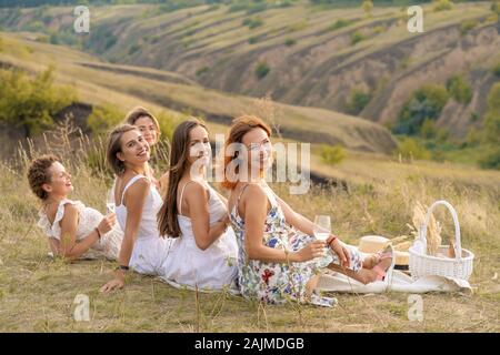 The company of cheerful female friends in white dresses enjoys a view of the green hills, relaxing on a picnic