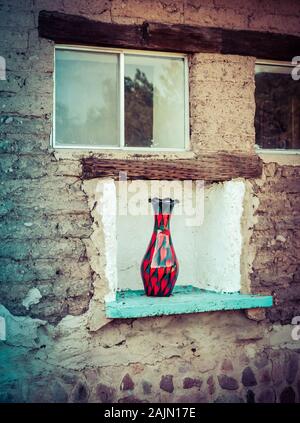 A curious site to see a contemporary red and black glass art vase sitting in an outdoor  wall niche on an old weathered, patched vintage adobe brick b Stock Photo