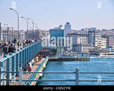 Galata bridge and Bosporus. Many people walking and fishing on the bridge under daylight. Residential buildings and metal railings. Stock Photo