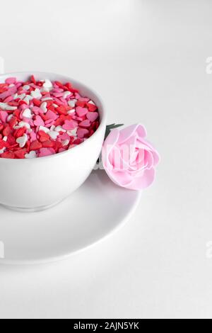white cup of coffee on a white background filled with heart shaped confetti. pink rose lies on the saucer selective focus. valentine day concept Stock Photo