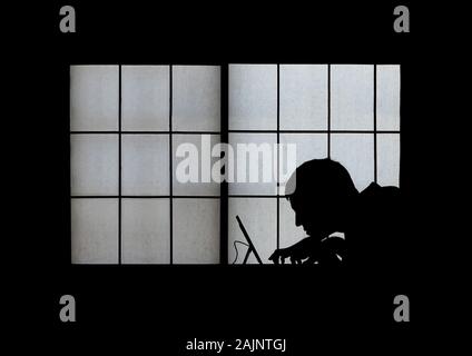 Window silhouette of a creepy computer hacker hunched over a laptop late at night Stock Photo