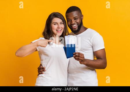 Honeymoon Tour Concept. Smiling interracial couple holding passports with boarding passes, ready to vacation travel, yellow background with copy space Stock Photo