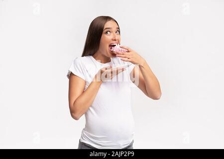 Pregnancy Diet. Hungry Pregnant Woman Biting Donut Standing On White Background. Studio Shot Stock Photo